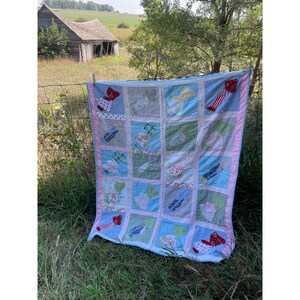 May include: A blue and pink patchwork quilt with appliqued red and white sock designs. The quilt is hanging on a clothesline in front of a wooden fence and a field.