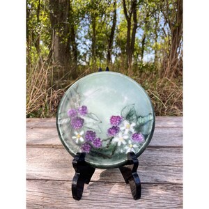 May include: A round, light green decorative plate with a hand-painted design of purple blackberries and white flowers. The plate is displayed on a black stand, set against a natural outdoor background.
