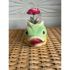 May include: A ceramic fish-shaped vase in a light green glaze with prominent dark red lips and black eyes. The vase features a wide opening at the top, currently holding a single artificial dark pink flower with green leaves and a small bud.