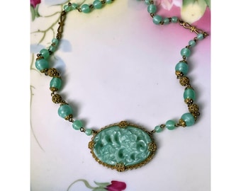 May include: A vintage green glass necklace with a carved oval pendant and gold tone metal accents. The necklace is made of round beads and has a clasp closure.