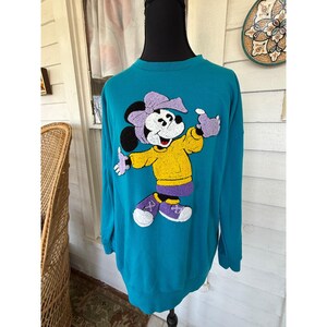 May include: Teal sweatshirt featuring a cartoon character with a purple bow, gloves, and skirt, and a yellow shirt. The character is wearing purple and white shoes. The sweatshirt is displayed on a black mannequin.