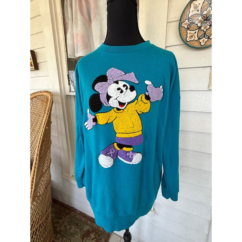 May include: Teal sweatshirt featuring a cartoon character with a purple bow, gloves, and skirt, and a yellow shirt. The character is wearing purple and white shoes. The sweatshirt is displayed on a black mannequin.