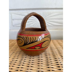 May include: A small, round, brown wooden basket with a handle. The basket is decorated with a tan design, black swirls, and red accents. The basket is sitting on a woven surface.