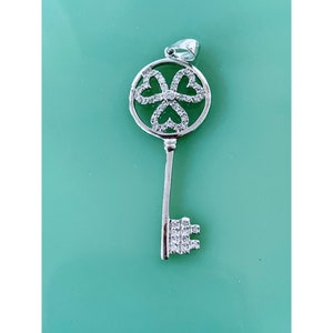 May include: A silver key pendant with a circular design featuring a four-leaf clover with hearts in the center. The pendant is adorned with clear cubic zirconia stones.