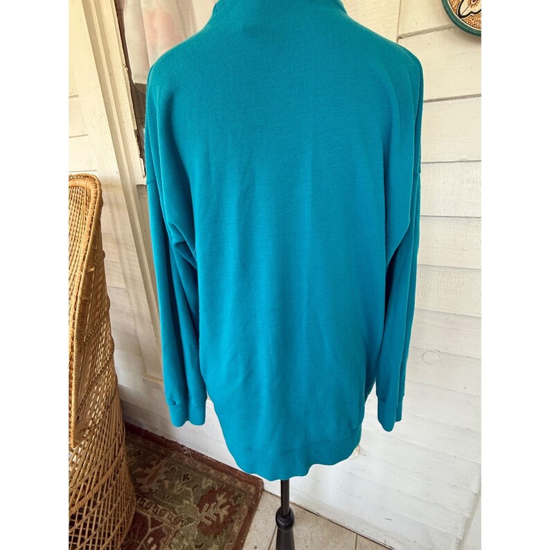 May include: Teal long-sleeved sweater with a mock neck. The back view of the sweater is displayed on a black stand. The sweater is made of a soft, knit material. The background includes a wicker chair, a rug, and a white wooden wall.