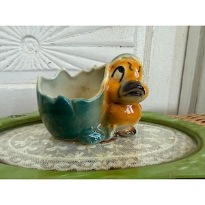 May include: A ceramic planter shaped like a yellow duckling emerging from a cracked eggshell. The eggshell portion is finished in a glossy teal glaze, while the duckling features painted black eyes and a dark beak, creating a hollow vessel.