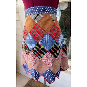 May include: A patchwork apron with a blue, red, brown, and black color scheme. The apron has a scalloped edge and a white polka dot trim.