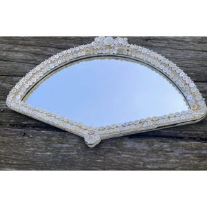 May include: A fan-shaped mirror with a white frame, intricately decorated with numerous small, off-white buttons. The mirror's reflective surface is smooth, and the buttons are arranged around the edges, creating a unique, handcrafted design.