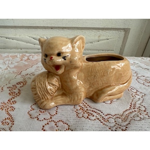 May include: A ceramic planter shaped like a cat holding a ball of yarn. The cat is light brown with black eyes and a red nose. The planter has a small opening on the top for planting.