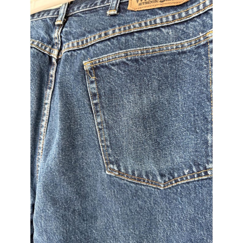 Puede incluir: Vaqueros de mezclilla azul oscuro con un dise&ntilde;o cl&aacute;sico de cinco bolsillos y costuras doradas visibles. En la cintura hay una etiqueta de cuero beige con el texto Wrangler Authentic Jeans impreso en tinta negra sobre la superficie.
