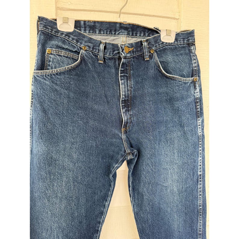 Puede incluir: Un par de pantalones vaqueros de color azul oscuro con un dise&ntilde;o cl&aacute;sico de cinco bolsillos, cierre de bot&oacute;n met&aacute;lico en la cintura, cremallera y trabillas resistentes para el cintur&oacute;n. La tela muestra un tono &iacute;ndigo lavado uniforme.