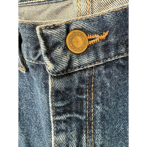 Puede incluir: Unos vaqueros de mezclilla azul con un bot&oacute;n de metal color lat&oacute;n que tiene grabada la palabra Wrangler. Los vaqueros est&aacute;n confeccionados con costuras gruesas de color naranja en contraste a lo largo de la bragueta y la cintura.