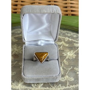 May include: A silver ring featuring a triangular tiger's eye gemstone with brown and gold bands, set in a polished metal band. The ring is displayed inside a grey velvet-lined jewelry box with a white satin interior lining and a hinged lid.