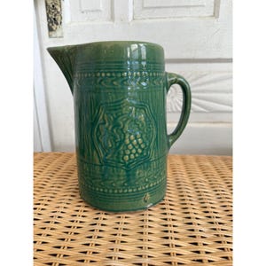 May include: Green ceramic pitcher with a spout and handle. The pitcher features embossed grape cluster and leaf designs, with decorative bands around the top and bottom. The pitcher is sitting on a wicker surface.