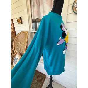 May include: Teal sweatshirt with a Minnie Mouse design. The long-sleeved top has a crew neck and a cartoon image of Minnie Mouse in purple, yellow, and white. The sweatshirt is displayed on a mannequin.