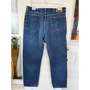 Puede incluir: Unos vaqueros de mezclilla azul oscuro con un dise&ntilde;o cl&aacute;sico de cinco bolsillos, cierre de bot&oacute;n y cremallera, y un parche de cuero marr&oacute;n en la cintura trasera con el nombre de la marca Wrangler impreso en texto negro.