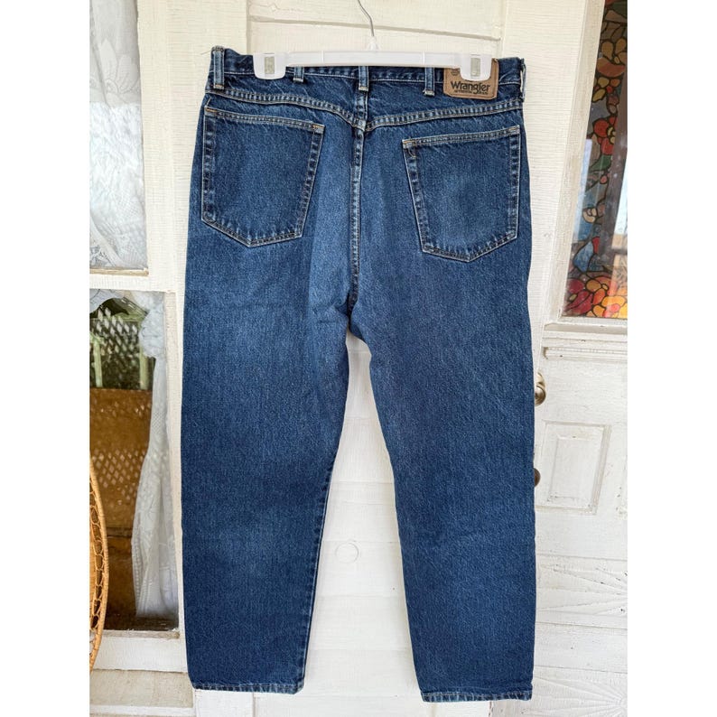 Puede incluir: Unos vaqueros de mezclilla azul oscuro con un dise&ntilde;o cl&aacute;sico de cinco bolsillos, cierre de bot&oacute;n y cremallera, y un parche de cuero marr&oacute;n en la cintura trasera con el nombre de la marca Wrangler impreso en texto negro.