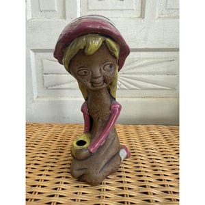 May include: A ceramic figurine of a seated figure with a whimsical design. The figure has a brown body, yellow hair, and a pink hat and sleeves. It holds a yellow object with a black opening. The figurine is on a woven surface.