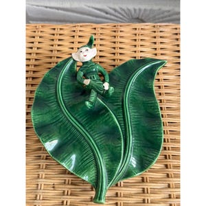 May include: A glossy ceramic dish shaped like two overlapping dark green leaves with a small, light-skinned elf figure wearing a green outfit and pointed hat sitting on the surface. The dish features detailed vein textures and a smooth finish.