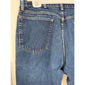 Puede incluir: Un par de pantalones vaqueros de mezclilla azul oscuro con un dise&ntilde;o de cintura alta, un bolsillo trasero visible, trabillas para cintur&oacute;n y costuras reforzadas a lo largo de los bordes. La tela tiene un lavado &iacute;ndigo cl&aacute;sico y textura.