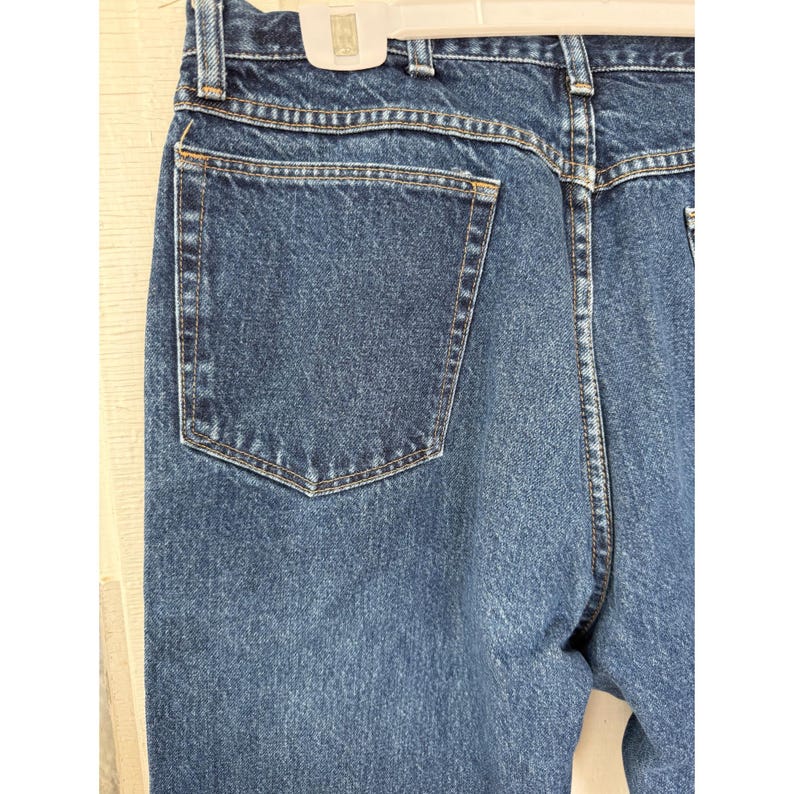 Puede incluir: Un par de pantalones vaqueros de mezclilla azul oscuro con un dise&ntilde;o de cintura alta, un bolsillo trasero visible, trabillas para cintur&oacute;n y costuras reforzadas a lo largo de los bordes. La tela tiene un lavado &iacute;ndigo cl&aacute;sico y textura.