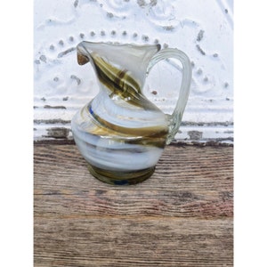 May include: A small, white glass pitcher with brown and blue swirl designs. The pitcher has a clear glass handle.