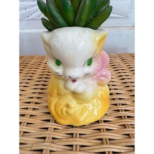May include: A vintage ceramic planter shaped like a cat, with a white head, yellow body, and pink bow. The cat has green eyes and a red mouth. The planter is filled with green succulent plants. The planter is sitting on a woven wicker surface.