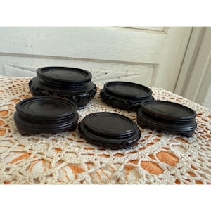 May include: Set of five black wooden round display stands of varying sizes. The stands are carved with intricate designs and are arranged on a white crocheted surface.