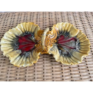 May include: A ceramic serving dish with two sections, each shaped like a scallop shell. The dish is decorated with a brown, yellow, and red glaze. The dish has a handle in the center.