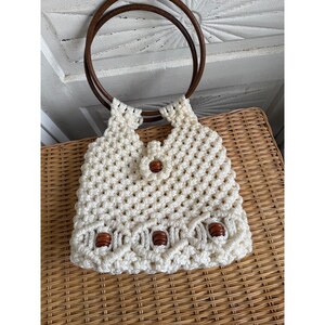 May include: A cream-colored macrame handbag featuring a knotted cord pattern, two circular dark brown wooden handles, and a central closure adorned with a single brown wooden bead. The lower section includes three additional brown wooden beads.