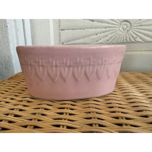 May include: A light pink ceramic bowl with a decorative pattern around the rim. The bowl has a slightly oval shape and sits on a woven wicker surface. The bowl's design features a repeating series of inverted teardrop shapes.