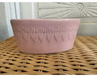 May include: A light pink ceramic bowl with a decorative pattern around the rim. The bowl has a slightly oval shape and sits on a woven wicker surface. The bowl's design features a repeating series of inverted teardrop shapes.