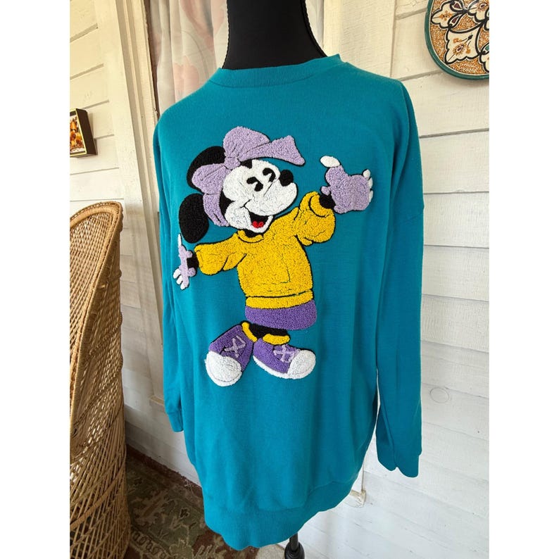 May include: Teal sweatshirt with an embroidered Minnie Mouse design. Minnie is depicted in a yellow top, purple shorts, and purple shoes. The long-sleeved, crew-neck sweatshirt is displayed on a mannequin.
