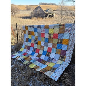 May include: A patchwork quilt made from a variety of fabrics in shades of blue, yellow, red, and brown. The quilt is hanging on a clothesline in front of a rustic barn.