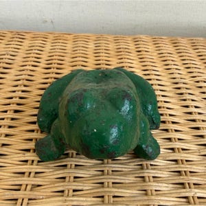 May include: A green frog-shaped cast iron doorstop. The doorstop has a textured surface and is painted in a distressed green color with dark spots. The frog is sitting on a woven wicker surface.