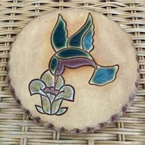 May include: Round ceramic coaster with a hummingbird design. The hummingbird is depicted in flight, with green, blue, and purple details. The coaster has a tan background and a flower design below the hummingbird.
