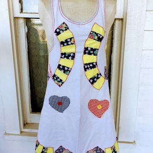 May include: A white apron with a patchwork design. The apron has yellow, black, red, blue, and white fabric. The apron has two heart-shaped patches with red and blue gingham fabric.