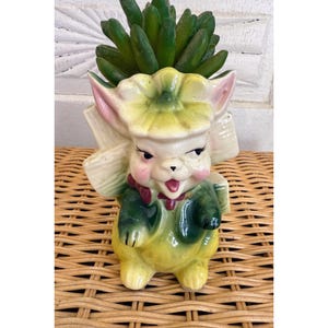 May include: A whimsical ceramic planter shaped like a cartoon character with a green and yellow outfit, pink cheeks, and a flower hat. The planter holds a cluster of green faux succulents. The overall design is vintage and decorative.