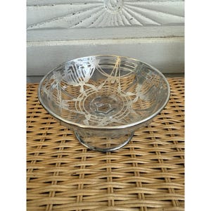May include: A clear glass bowl with etched floral designs and a silver-colored rim. The bowl sits on a silver-colored metal base. The bowl is on a woven wicker surface.