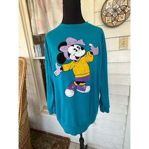 May include: Teal sweatshirt with a Minnie Mouse design. The cartoon character is wearing a yellow jacket, purple shoes, and a purple bow. The sweatshirt has long sleeves and a crew neck.