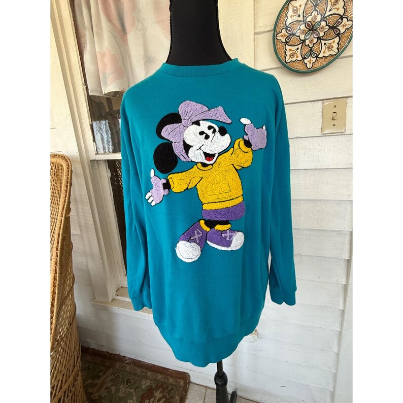 May include: Teal sweatshirt with a Minnie Mouse design. The cartoon character is wearing a yellow jacket, purple shoes, and a purple bow. The sweatshirt has long sleeves and a crew neck.