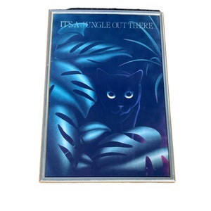 May include: A framed print of a black cat with blue eyes peeking out from behind large blue leaves. The print has the text "It's a jungle out there" at the top.