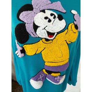 May include: Teal sweatshirt with a large, textured Minnie Mouse design. Minnie is wearing a yellow sweater, purple shoes, and a matching bow. The design has black, white, yellow, purple, and red accents. A fun, colourful, and playful design.