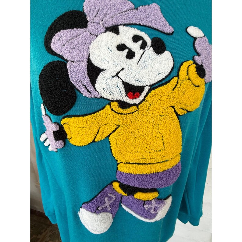 May include: Teal sweatshirt with a large, textured Minnie Mouse design. Minnie is wearing a yellow sweater, purple shoes, and a matching bow. The design has black, white, yellow, purple, and red accents. A fun, colourful, and playful design.
