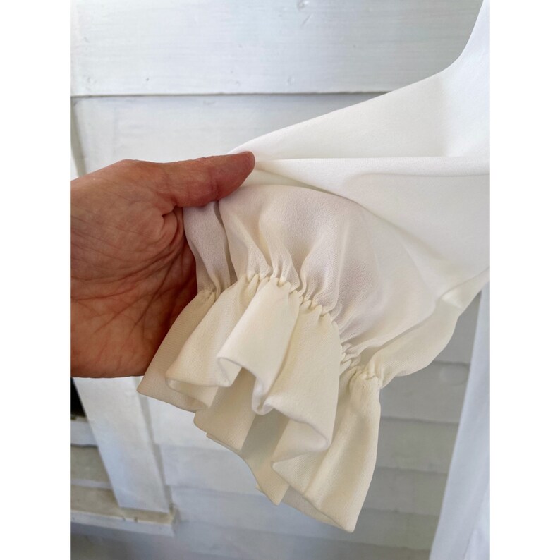 Op de afbeelding: Close-up van een wit, gerimpeld mouwdetail op een kledingstuk. De stof is zacht en soepel, met een delicate, geplooide look.