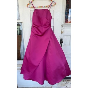 May include: A fuchsia-colored formal dress with a draped, asymmetrical skirt. The dress features a fitted bodice with spaghetti straps and a ruched detail on the side. The dress is hanging on a colorful beaded hanger.