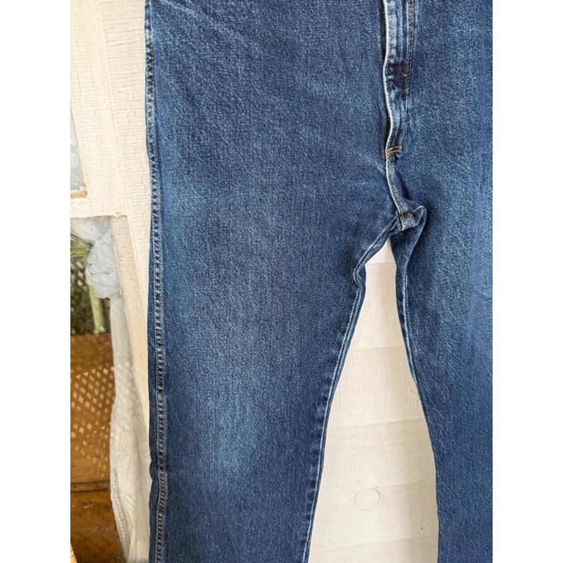Puede incluir: Unos pantalones vaqueros de color azul oscuro con un dise&ntilde;o cl&aacute;sico de cinco bolsillos, cierre de cremallera con bot&oacute;n met&aacute;lico y costuras naranjas visibles a lo largo de las costuras. La tela tiene una textura de lavado medio uniforme.
