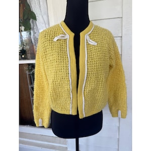 May include: A vintage yellow knit cardigan with a textured pattern. The cardigan features white trim along the collar, front edges, and cuffs, with small bow accents. The open front design is displayed on a black mannequin.