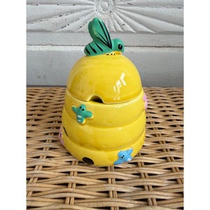 May include: A yellow ceramic honey pot shaped like a beehive, with a green and black bee on top. The pot has a small opening for a spoon and is decorated with small, colorful bees.