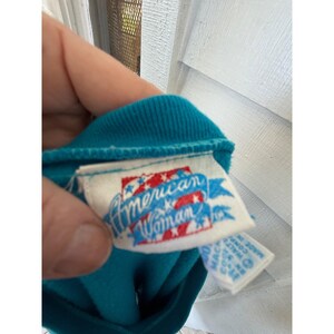 May include: A teal garment with a white label featuring the text "American Woman". The label has red and blue stars and stripes. The garment's collar is visible. The image is a close-up.
